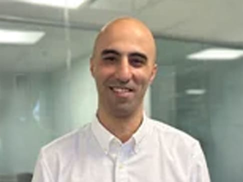 A smiling bald man wearing a white button-up shirt stands in an office setting with glass walls in the background.