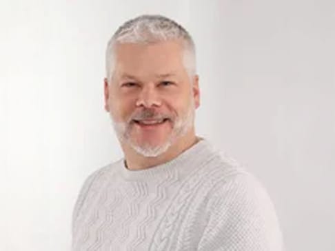 Smiling man with gray hair and a beard, wearing a white textured sweater, stands against a light background.