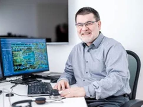 A middle-aged man with glasses smiles while sitting at a desk. He is working on a computer with a colorful screen displaying digital graphics. There are headphones and a mouse on the desk.