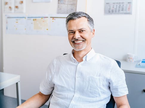 A smiling middle-aged man with a short beard and gray hair sits in an office, wearing a white button-up shirt. Behind him are papers and calendars on the wall.