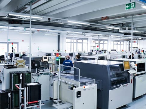 A spacious manufacturing facility filled with machines and workers. Several high-tech machines are lined up on the factory floor, and employees can be seen engaged in various tasks. Large windows allow natural light to fill the room, and safety signs are visible.