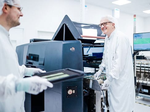 Two men in white lab coats and hairnets work together in a printing facility. One man is smiling and engaged with the machinery, while the other is holding a printing plate. High-tech equipment and screens are visible in the background.