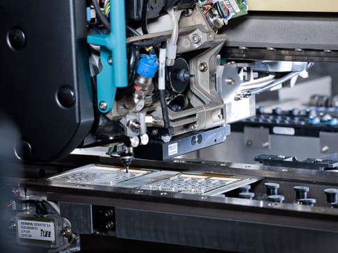 Eine Nahaufnahme einer automatisierten Maschine, die an einer Leiterplatte arbeitet. Verschiedene mechanische Teile und Kabel sind sichtbar, während die Maschine präzise Bewegungen ausführt.