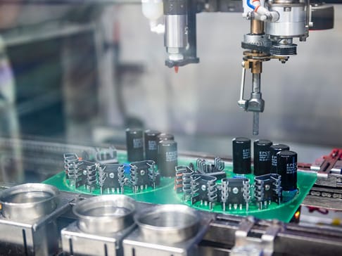 Roboterarm montiert elektronische Bauteile auf einer Leiterplatte in einer modernen Fertigungseinrichtung. Die Szene zeigt verschiedene Kondensatoren und Bauelemente auf einem Förderband.