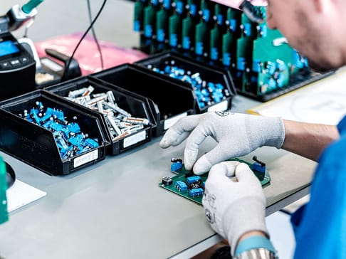 Ein Arbeiter in Handschuhen montiert Bauteile auf einer Leiterplatte. Im Hintergrund befinden sich mehrere Schalen mit ähnlichen Bauteilen und Werkzeugen zum Testen.