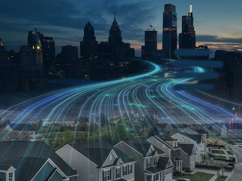 A skyline view of a city at dusk, featuring tall buildings and skyscrapers, with glowing blue and green lines flowing through the air, suggesting data movement or connectivity over residential neighborhoods in the foreground.