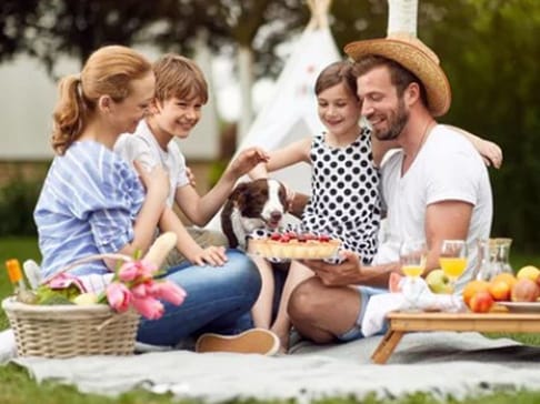 A family picnic scene featuring four people sitting together on a blanket. Two children are petting a dog, while the adults are smiling and enjoying a cake. A basket of flowers and fruits is nearby. The setting is outdoor with a teepee in the background.