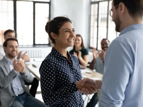 A woman with dark hair smiles while shaking hands with a man in front of a group of people applauding in a bright, modern meeting room.