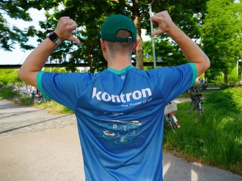A person wearing a blue athletic shirt with the word "kontron" and the phrase "The Power of IoT" on the back stands outdoors, flexing their arms in a celebratory pose. Lush greenery surrounds them, indicating a pleasant day.