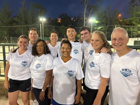 A group of eight people, wearing matching white t-shirts with "Kontron" printed on them, pose for a photo on a sports field at dusk. They are smiling and appear to be friends or colleagues. Trees and lights are visible in the background.