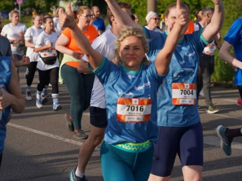 A group of diverse runners participating in a race, with one woman in the foreground smiling and giving a thumbs-up. Most runners wear matching blue and orange shirts, with bib numbers visible on their chests. The scene is lively and energetic, set along a bright, sunny path.
