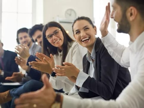 A group of six people seated in a bright room, all smiling and clapping. One woman in the foreground, wearing a blazer, is laughing, while others are engaged and supportive.