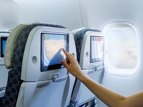 飛行機の座席の背もたれにあるタッチスクリーンを操作する手。窓の外には雲が見える。