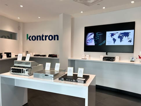 A modern display area showcases various electronic devices and equipment with a Kontron logo on the wall. A large screen features a map and an eye graphic, enhancing the tech-themed environment. Labels describe each device on the white tables.