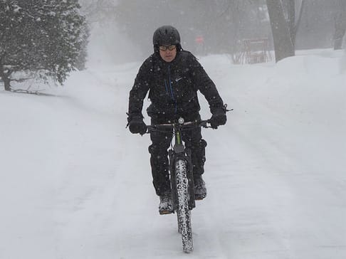 A cyclist dressed in black winter gear rides a bicycle on a snow-covered path during a blizzard, with snowflakes swirling around and visibility reduced.