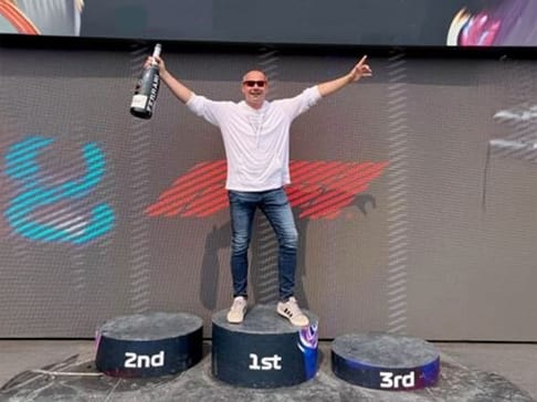 Un homme se tient sur un podium, levant un flacon dans une main et faisant un signe de victoire avec l'autre. Il est au centre du podium, avec des numéros indiquant 1er, 2e et 3e.