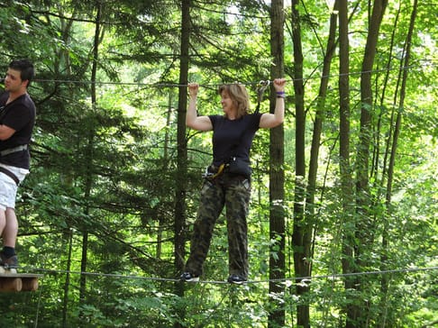 Une femme en équilibre sur un câble, en pleine forêt, tandis qu'un homme l'observe de près. Elle porte un équipement de sécurité et des vêtements de camouflage, entourée de verdure.