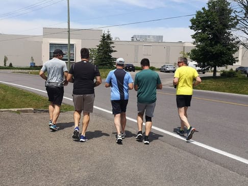 Un groupe de cinq hommes en train de courir sur un chemin, tournés vers l'arrière. Ils portent des vêtements de sport variés et progressent sur une route bordée d'arbres et de bâtiments.