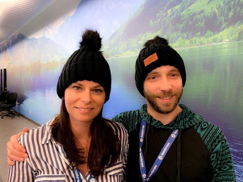 Deux personnes souriantes portent des bonnets noirs avec un pompon. L'une est en chemise rayée, tandis que l'autre porte un pull à motifs. En arrière-plan, un paysage de montagnes et d'eau.