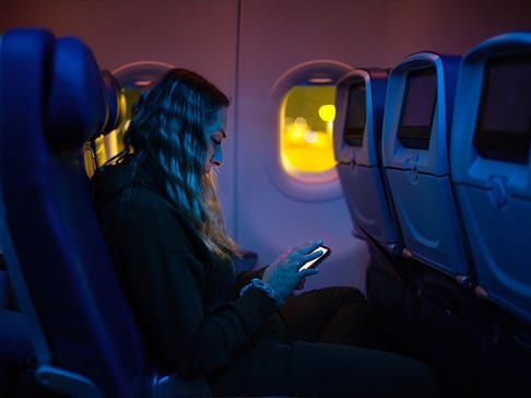 A woman with wavy hair sits in an airplane seat, illuminated by the glow of her phone, surrounded by dark blue hues. The cabin is quiet, with empty seats next to her and a soft light from outside the window.