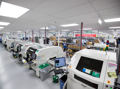 Usine moderne avec plusieurs machines de production électronique. Des employés surveillent le processus de fabrication, tandis que des boîtes sont visibles sur des étagères. L'éclairage est fluorescent et le sol est propre.