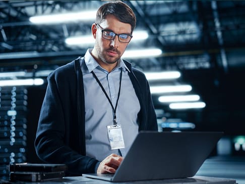 Un homme en lunettes et pull, travaillant sur un ordinateur portable dans un environnement industriel éclairé. Il porte un badge d'identification.