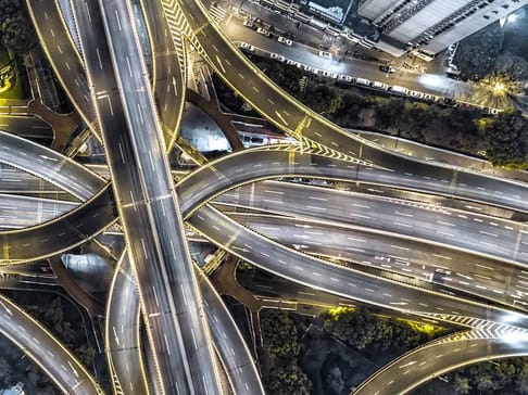 Ein komplexes Autobahnkreuz mit mehreren Überführungen und Fahrbahnen, beleuchtet von Scheinwerfern in der Nacht.