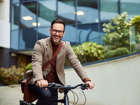 Ein lachender Mann in einer braunen Jacke und Brille fährt mit einem Fahrrad durch eine moderne urbane Umgebung. Pflanzen und Glasarchitektur sind im Hintergrund sichtbar.