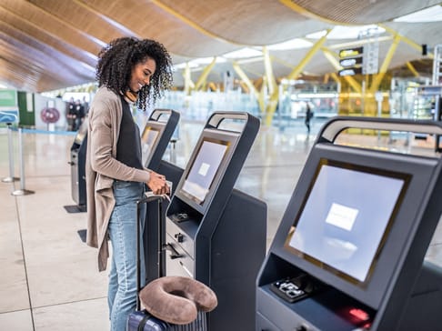 Eine Person mit lockigem Haar steht an einem Selbstbedienungscheck-in-Terminal am Flughafen. Sie hat einen Koffer bei sich und scheint dabei, ihre Reise zu beginnen. Der Flughafen hat eine moderne Architektur mit vielen Passagieren im Hintergrund.