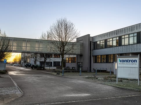 Ein moderner Gebäudekomplex mit einer Verbindung aus Glas im Vordergrund. Der Schriftzug „kontron Technologie-Campus“ ist gut sichtbar. Bäume und Parkplätze umgeben das Gebäude, während die Morgensonne den Himmel beleuchtet.