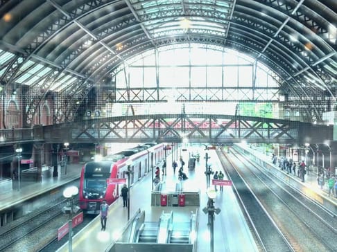 Ein belebter Bahnhof mit einem roten Zug, der an einem Gleis steht. Menschen warten oder bewegen sich in verschiedene Richtungen. Die Architektur des Daches ist beeindruckend mit großen Fenstern und metallischen Strukturen.