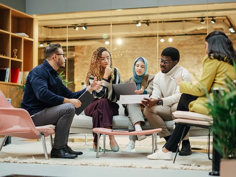 Fünf Personen sitzen in einem modernen Raum auf einem Sofa und diskutieren, während sie Laptops und Notizen verwenden. Die Gruppe besteht aus verschiedenen ethnischen Hintergründen und trägt legere Kleidung.