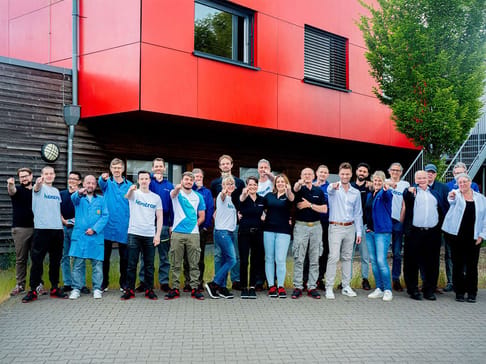 A diverse group of about 30 people stands together in front of a red and wooden building, smiling and posing for the camera. They wear a mix of casual attire, some featuring the logo "kreator." Trees and greenery are visible in the background.