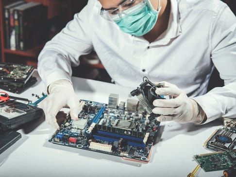 Eine Person mit medizinischer Maske und Handschuhen arbeitet konzentriert an einer Hauptplatine. Um sie herum liegen verschiedene Computerbauteile und Werkzeuge.