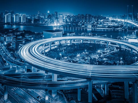 Ein nächtlicher Blick auf eine Stadt mit mehrstöckigen Autobahntrassen und leuchtenden Lichtern. Im Hintergrund erstreckt sich die Skyline mit hochaufragenden Gebäuden entlang eines Flusses.