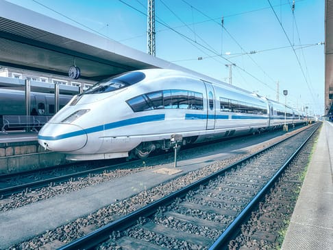 A sleek, modern high-speed train sits at a railway station platform under a clear blue sky. The train features a silver body with blue accents and is positioned on the tracks with station infrastructure visible in the background.