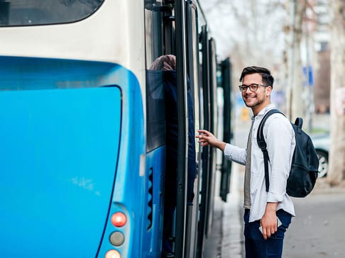 A young man with glasses and a backpack smiles as he prepares to board a blue bus. The background features trees and a city street.