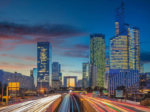 A vibrant city skyline at dusk, showcasing tall skyscrapers with illuminated windows. Traffic trails light up the road below, while the sky transitions from orange to deep blue.