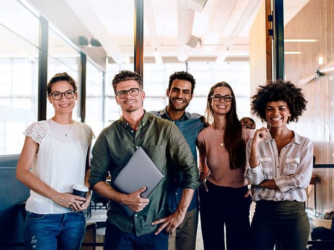 A diverse group of five smiling individuals stands together in a modern office setting. They are all wearing glasses, with three men and two women, and display a mix of casual and professional attire. One person holds a laptop, while another has a coffee cup. The background features large windows and wood paneling.