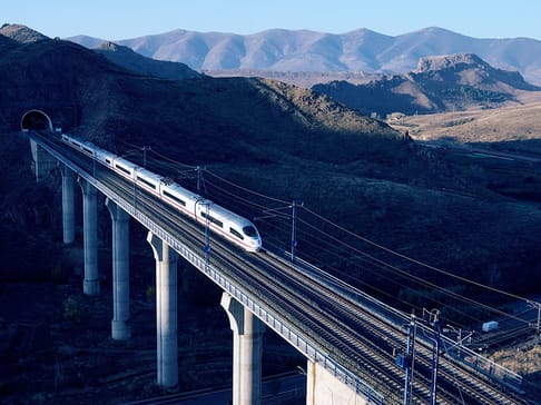 A high-speed train travels along a viaduct in a mountainous landscape, with blue sky and distant hills in the background.