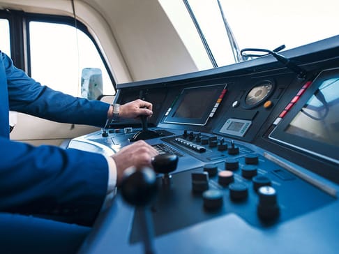 A person in a suit is operating a vehicle's control panel, featuring various buttons, levers, and screens. The interior is modern with large windows showing a bright sky.