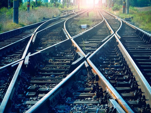 Railway tracks converge and diverge at a junction, with sunlight illuminating the distant end of the tracks. Grass and trees border the sides, creating a natural setting.