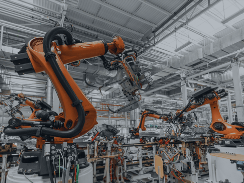 A factory setting featuring multiple orange robotic arms engaged in automated assembly tasks. The environment is spacious, with overhead lighting and various machinery in the background.