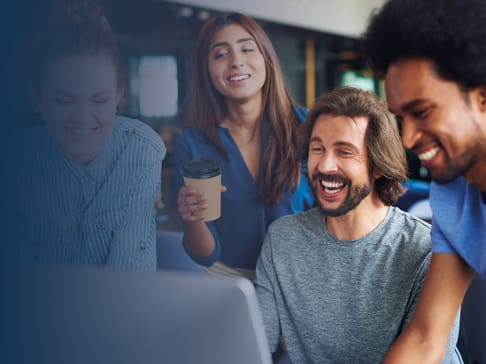 A group of four diverse friends are gathered around a laptop, laughing and enjoying each other's company. One person holds a coffee cup, while the others look intently at the screen. They appear engaged and happy in a casual, modern environment.