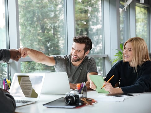A man and a woman share a friendly fist bump while sitting at a table with laptops and notebooks. The man is smiling and wearing a gray t-shirt, while the woman holds a green notepad and pencil, also smiling. Both are in a bright, modern workspace with large windows and greenery in the background.