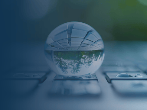 A crystal clear water droplet sits on a flat surface, reflecting a blurred view of greenery above and a tiled pattern below. Soft blue tones surround the droplet, creating a serene atmosphere.