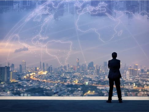 A person in a suit stands with their back to the viewer, gazing out at a city skyline illuminated by lights. A digital map overlay highlights global connections against a twilight sky filled with skyscrapers.