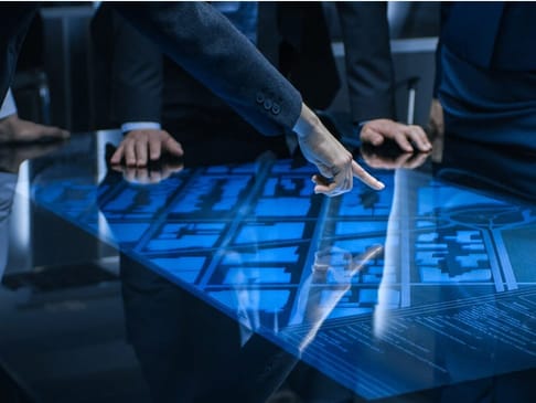 A person's hand points at a glowing, holographic map on a table, while others lean in to examine the display. The environment appears modern and high-tech, with a focus on the digital map's detailed layout.