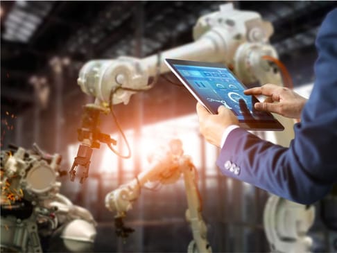A person in a suit interacts with a tablet displaying control options while overseeing robotic arms in a factory setting. Sparks and industrial elements suggest an active manufacturing environment.
