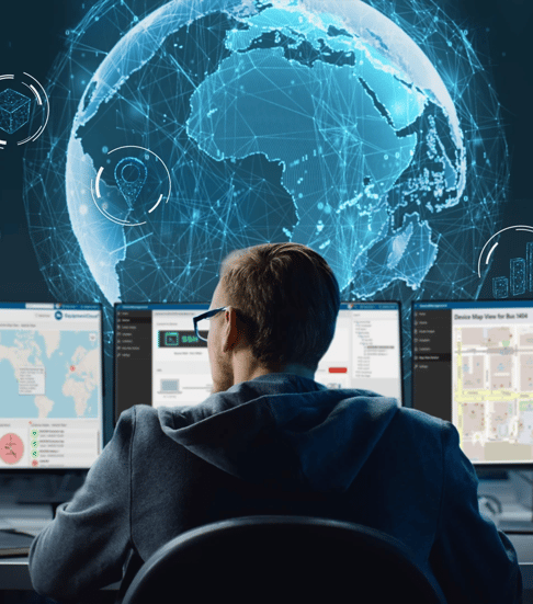 A person sits at a desk in front of multiple computer screens displaying maps and data visualizations, with a digital globe and network graphics projected in the background.